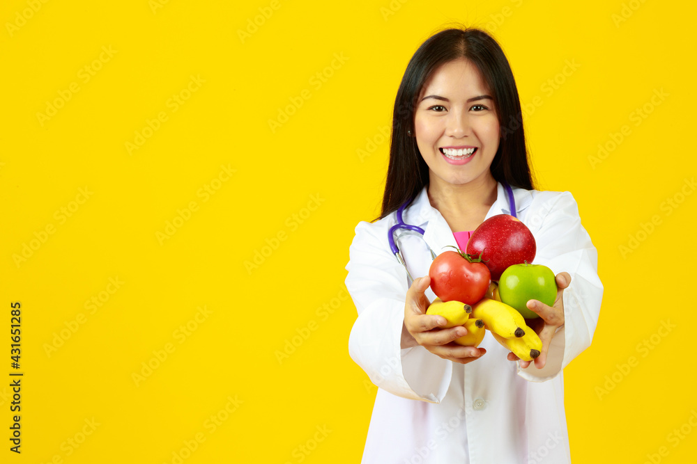 Asian young smiley female doctor wearing white gown uniform with stethoscope, holding banana, apples and tomato, presenting fruit vitamin, healthy with isolated yellow background and copy space. Asian young smiley female doctor wearing white gown uniform with stethoscope, holding banana, apples and tomato, presenting fruit vitamin, healthy with isolated yellow background and copy space.