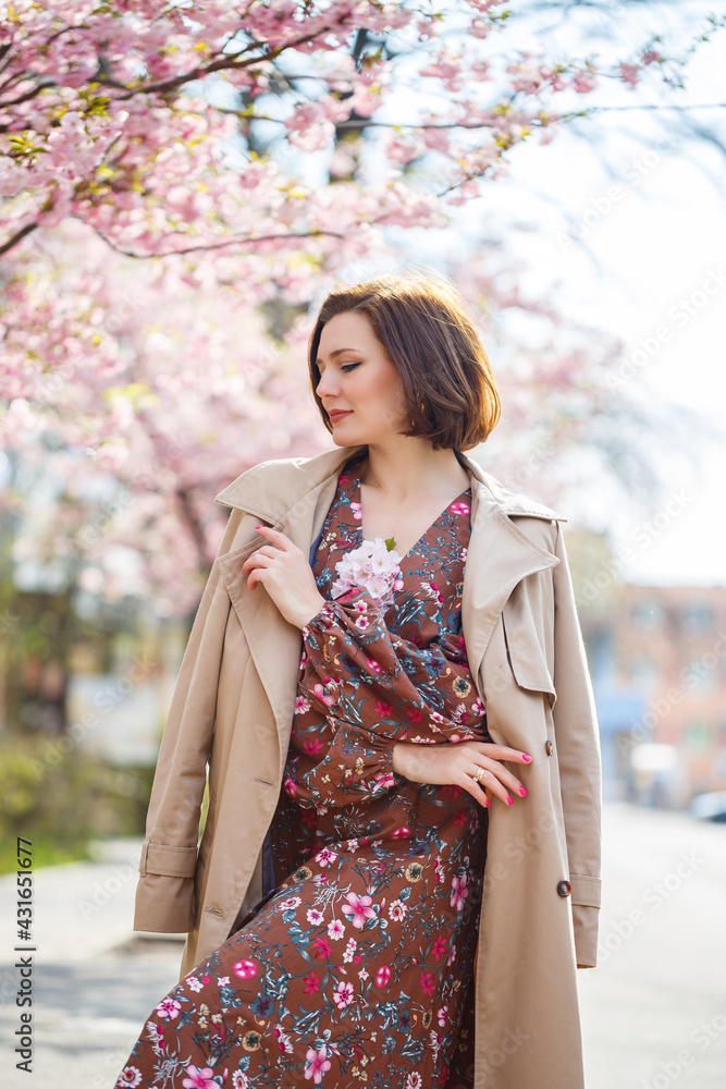 Portrait of a gentle woman against the background of sakura flowers. Walk in the blossoming sakura garden. Young stylish woman standing in sakura park and enjoying beauty Portrait of a gentle woman against the background of sakura flowers. Walk in the blossoming sakura garden. Young stylish woman standing in sakura park and enjoying beauty