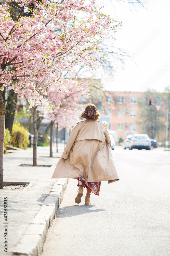 In the spring, a woman walks along a blooming street with sakura trees. A girl in a long silk elegant vintage dress walks among the flowering trees