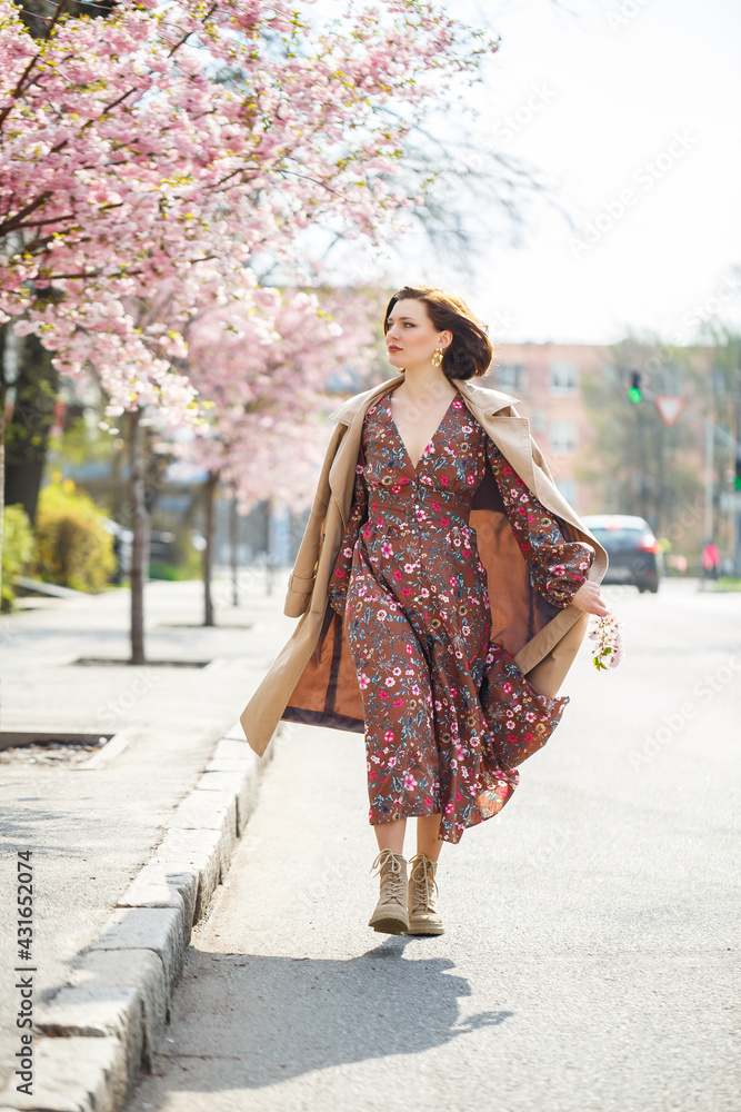 In the spring, a woman walks along a blooming street with sakura trees. A girl in a long silk elegant vintage dress walks among the flowering trees In the spring, a woman walks along a blooming street with sakura trees. A girl in a long silk elegant vintage dress walks among the flowering trees