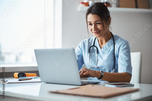 Leinwand Poster Joyful female doctor using notebook in clinic