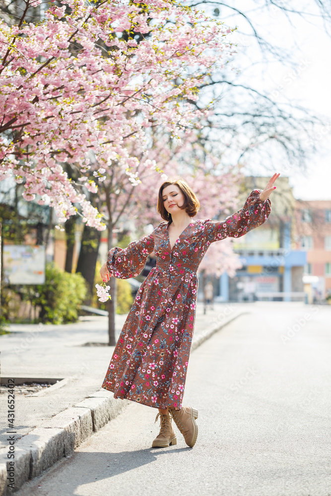 In the spring, a woman walks along a blooming street with sakura trees. A girl in a long silk elegant vintage dress walks among the flowering trees