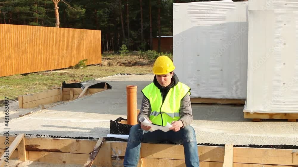 Construction worker in a protective yellow hardhat and a signal vest sit down in foundation with wooden formwork on a construction site and studies the drawings and documentation. Building materials. Construction worker in a protective yellow hardhat and a signal vest sit down in foundation with wooden formwork on a construction site and studies the drawings and documentation. Building materials.
