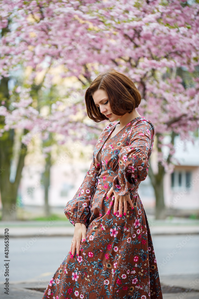 A woman walks along a spring street. The girl enjoys the scent of a blossoming tree. Beautiful woman in a dress with blooming cherry sakura