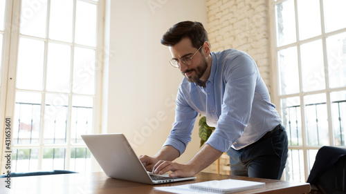 Happy young male manager employee in eyeglasses working on computer standing at table, typing email, communicating distantly with clients or colleagues or preparing electronic research report.