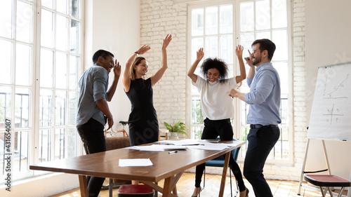 Overjoyed millennial diverse mixed race business people dancing in modern office, celebrating shared corporate success. Emotional happy multiracial employees having fun, relaxing together at workplace