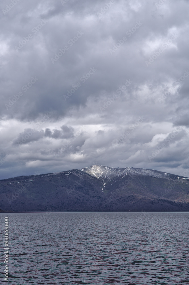 波立つ湖の向こうに見る曇り空の下の残雪の山。