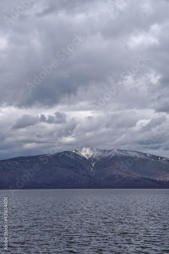 波立つ湖の向こうに見る曇り空の下の残雪の山。