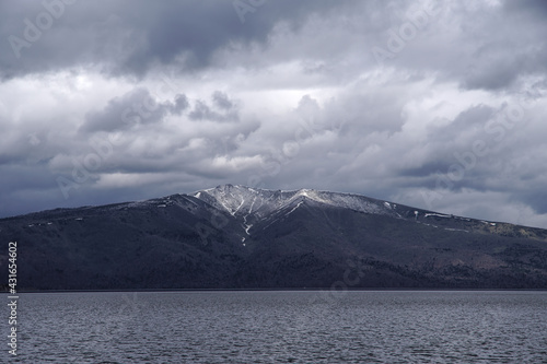 波立つ湖の向こうに見る曇り空の下の残雪の山。 © Masa Tsuchiya