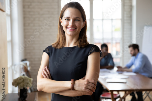 Portrait of confident smiling attractive female team leader in formal dress standing in modern workplace. Happy young 30s businesswoman boss employer partner posing indoors, looking at camera.