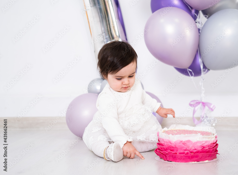 baby girl in white dress sitting on the floor celebrating her first birthday with cake and balloons.