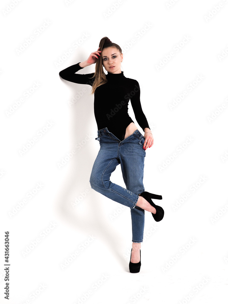 The brunette stands on a white background in a black bodysuit, jeans and high heels The brunette stands on a white background in a black bodysuit, jeans and high heels