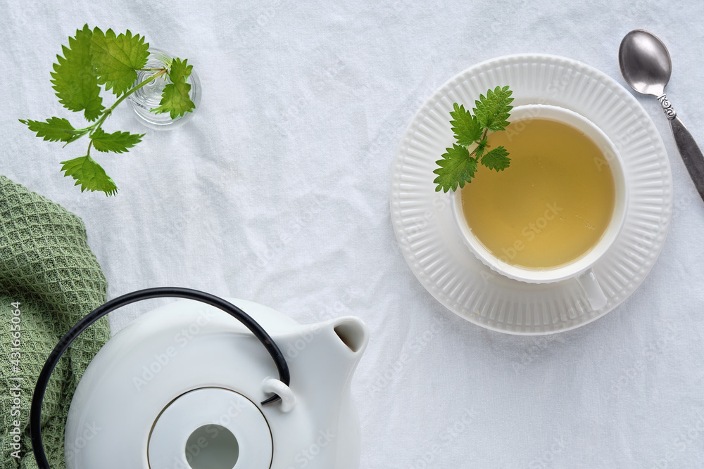 Nettle tea cup, fresh stinging nettle herbs. Flat lay, top view, off white textile tablecloth. White ceramic teapot. Natural light. - obrazy, fototapety, plakaty 