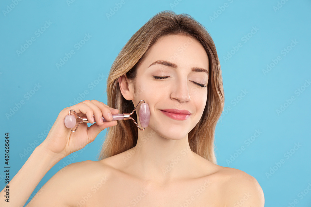 Young woman using natural rose quartz face roller on light blue background Young woman using natural rose quartz face roller on light blue background