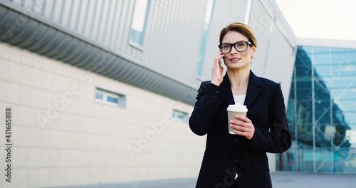 Attractive smiled woman boss walking on the modern office center background, talking on the phone and holding her morning coffee on a sunny day. Outdoors