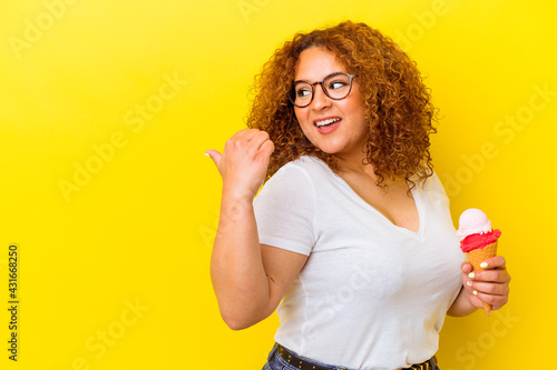 Young latin woman holding an ice cream isolated on yellow background points with thumb finger away, laughing and carefree.