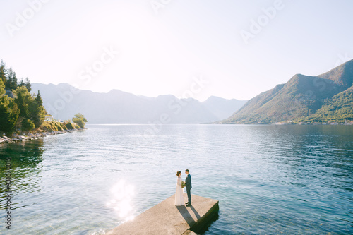 Newlyweds stand on the pier holding hands on a sunny warm day against the backdrop of mountains