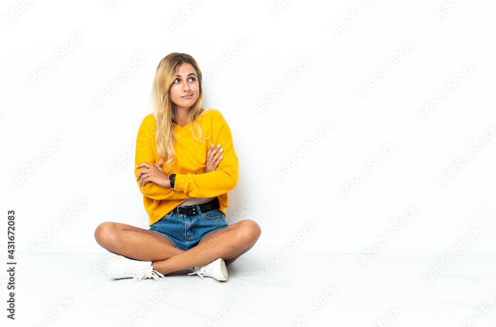 Young blonde Uruguayan woman sitting on the floor isolated on white background making doubts gesture while lifting the shoulders