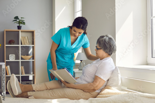Female home care nurse assists old woman with daily needs. Caregiver for elderly patients talking to relaxed senior lady who\'s sitting on comfortable bed at home, reading book and enjoying free time