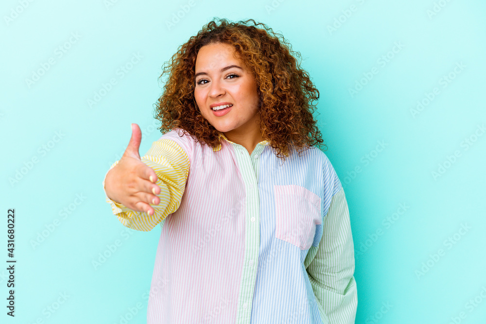 Young latin curvy woman isolated on blue background stretching hand at camera in greeting gesture. Young latin curvy woman isolated on blue background stretching hand at camera in greeting gesture.