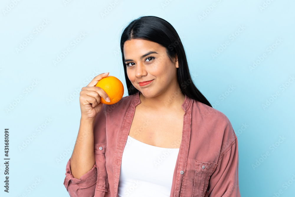 Young latin woman woman isolated on blue background holding an orange Young latin woman woman isolated on blue background holding an orange