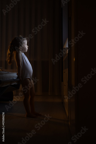 a little girl stands in a dark room and watches TV