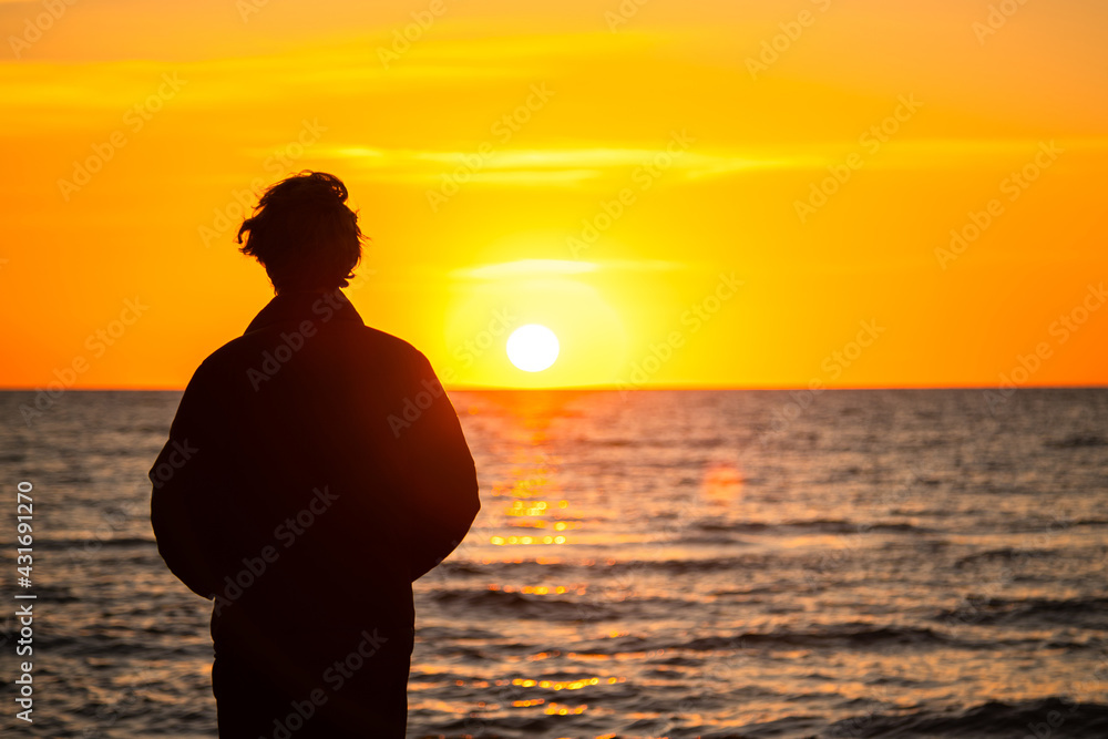 Dark silhouette of a man standing by the sea at golden sunset. Lonely teenager looking a setting sun.