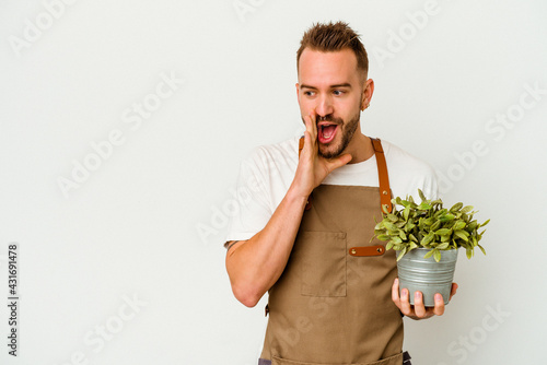 Young gardener tattooed caucasian man holding a plant isolated on white background is saying a secret hot braking news and looking aside