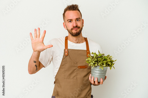 Young gardener tattooed caucasian man holding a plant isolated on white background smiling cheerful showing number five with fingers.