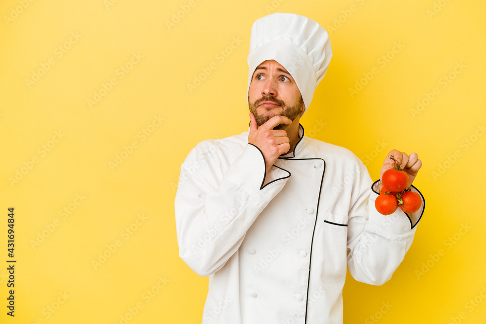 Young caucasian chef man holding tomatoes isolated on yellow background looking sideways with doubtful and skeptical expression. Young caucasian chef man holding tomatoes isolated on yellow background looking sideways with doubtful and skeptical expression.