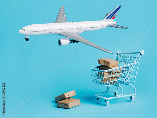 Plane flies around the shopping trolley and cardboard boxes on blue background. Creative cargo delivery by air concept. Free and fast freight global transportation.