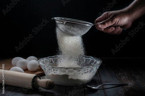 Sifting flour into a cristal bowl with eggs on the side and on a table, with a black background, bakery, or cooking concept dark food or low key light photography