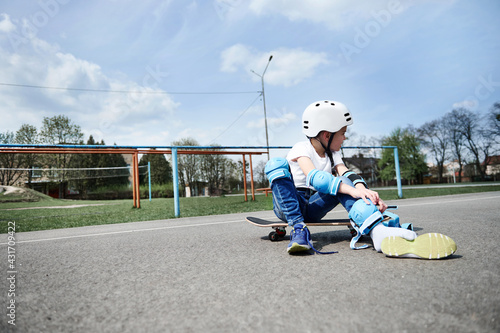 Child boy skateboarder looking away while sitting on a skateboard and putting on protective equipment