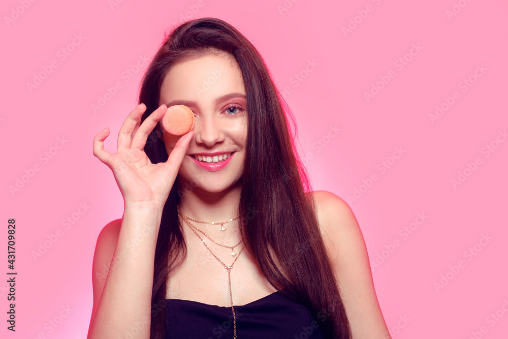 Fototapeta premium Girl eats macaroon, model on a pink background eats a sweet, sugar diet. Brunette with pink make-up, addicted to food. Smiles