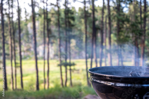 Focus on an open portable charcoal grill at a campsite overlooking a green forest