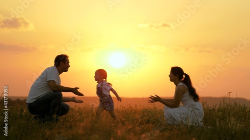 Happy family walks in park at sunset. Mom, dad and baby. Little daughter goes from mom to dad, hugs and kisses her parents in rays of a warm sun. Healthy family plays in field. Happy family concept