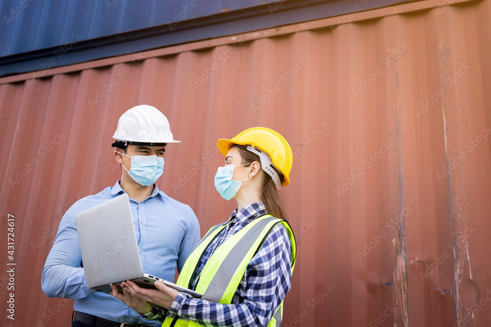 Team of logistic engineer man and woman wearing helmets with laptops holding hand and protective face mask standing outside containers area warehouse Team of logistic engineer man and woman wearing helmets with laptops holding hand and protective face mask standing outside containers area warehouse