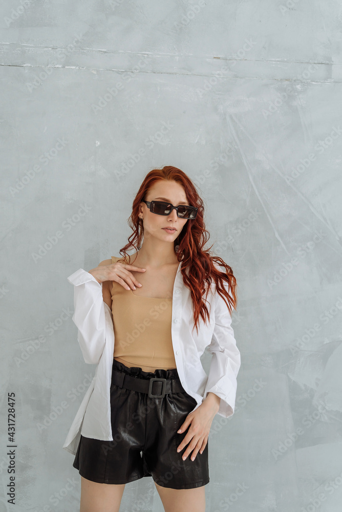 A fashionable red-haired beautiful female model wearing sunglasses, a large white shirt and black shorts. beautiful sexy woman perfect skinny body shape. clothing for the summer season A fashionable red-haired beautiful female model wearing sunglasses, a large white shirt and black shorts. beautiful sexy woman perfect skinny body shape. clothing for the summer season