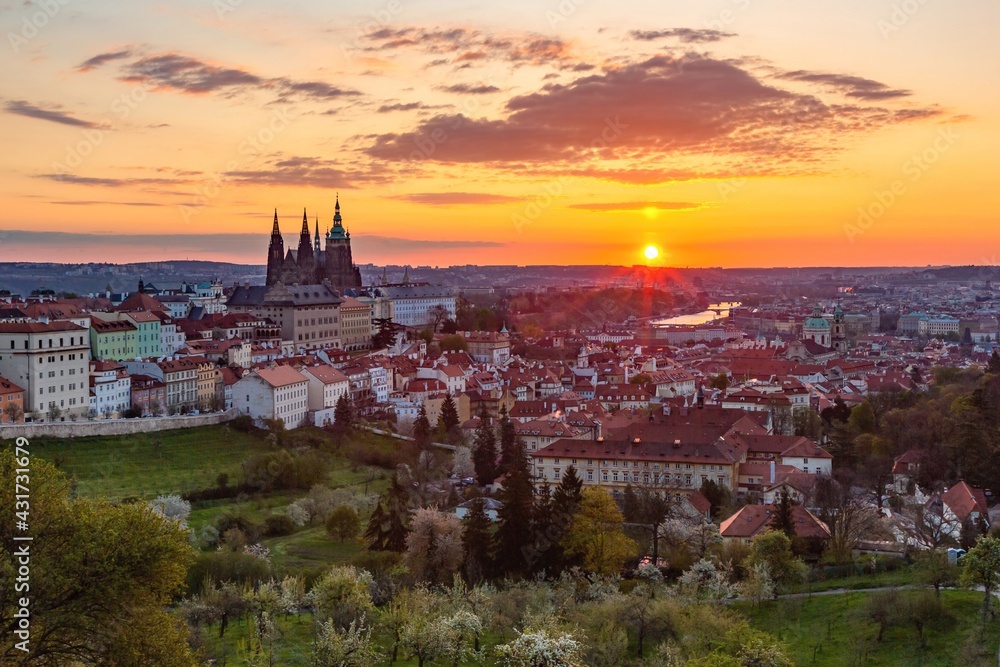 Prague, Czech Republic - May 3 2021: Colorful sunrise view of the city from Petrin hill with the castle, garden, trees, river and buildings. Clouds on the yellow, orange, pink and blue sky.