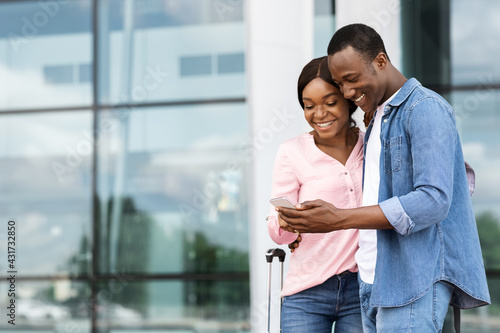 Cheerful black couple with smartphone booking hotel online, standing near arport building