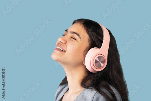 Music lover. Calm Indian teen girl in headphones listening to beautiful song with closed eyes on blue background