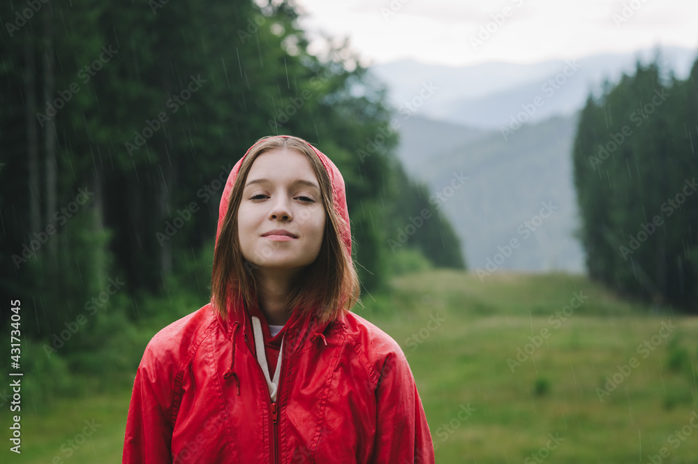 Girl in a red raincoat stands in mountains against the backdrop of a beautiful landscape and enjoys the rain, looks in camera and smiles. Positive female tourist has fallen in the rain in mountains. Girl in a red raincoat stands in mountains against the backdrop of a beautiful landscape and enjoys the rain, looks in camera and smiles. Positive female tourist has fallen in the rain in mountains.