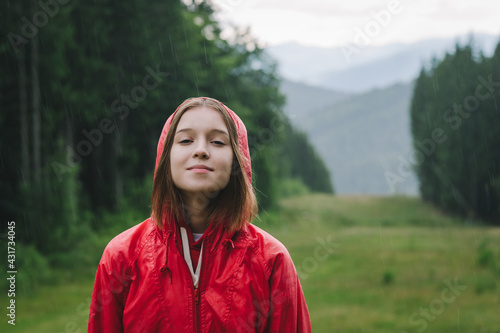 Girl in a red raincoat stands in mountains against the backdrop of a beautiful landscape and enjoys the rain, looks in camera and smiles. Positive female tourist has fallen in the rain in mountains.