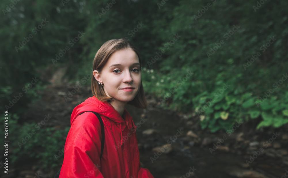 Closeup portrait of a smiling girl in a red raincoat hiking, looking into the camera and smiling against the backdrop of a mountain stream and pristine nature. Hiking in the mountain forest. Closeup portrait of a smiling girl in a red raincoat hiking, looking into the camera and smiling against the backdrop of a mountain stream and pristine nature. Hiking in the mountain forest.