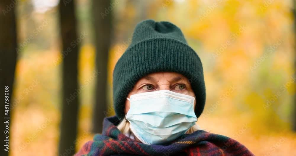 Woman 50s, wearing warm clothes and a protective medical mask, looks at the camera. Walking in a park in an autumn forest. Woman 50s, wearing warm clothes and a protective medical mask, looks at the camera. Walking in a park in an autumn forest.