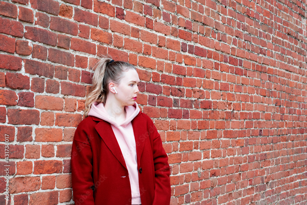Young woman wearing earbuds, woolen coat and hoodie is standing by the brick wall and is looking away. Copy space Young woman wearing earbuds, woolen coat and hoodie is standing by the brick wall and is looking away. Copy space