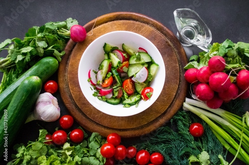 A pile of fresh, ripe vegetables: radishes, tomatoes, cucumbers, herbs, and garlic are arranged around a round wooden chopping board. A white bowl with vegetables cut into a salad and olive oil