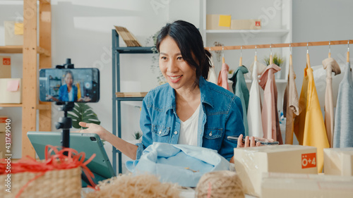 Obraz na plátně Young Asia lady fashion designer using mobile phone receiving purchase order and showing clothes recording video live streaming online at shop
