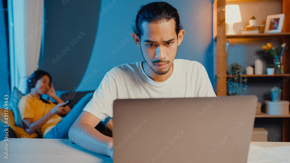 Freelance asian couple man and woman in casual husband focus work in laptop computer at night wife take a rest feel relax wear headphones play music on smartphone lay down and dance on couch at home. Freelance asian couple man and woman in casual husband focus work in laptop computer at night wife take a rest feel relax wear headphones play music on smartphone lay down and dance on couch at home.