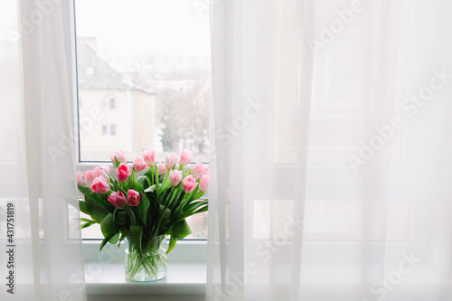 Bouquet of pink tulips in transparent vase on windowsill  against background of window. Flowers in interior.